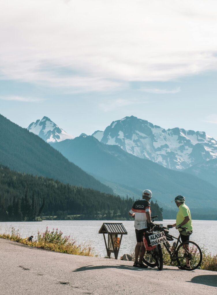 Two people on bikes near a lake with mountains in the background